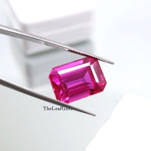 Puede incluir: Una gema rosa brillante, rectangular y facetada, es sostenida por unas pinzas plateadas sobre un fondo blanco. La gema tiene una superficie brillante y reflectante. El texto "TheLeafGifts" es visible en las pinzas.
