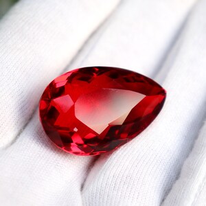 May include: A faceted, teardrop-shaped gemstone in a vibrant red hue. The gem's cut reveals multiple internal facets, creating a play of light and shadow. The stone is displayed against a white background.
