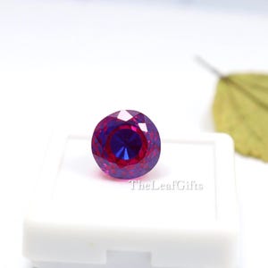 May include: A round, faceted gemstone with a colour gradient from deep magenta to royal blue. The gem is displayed in a white case, with the text "TheLeafGifts" visible.