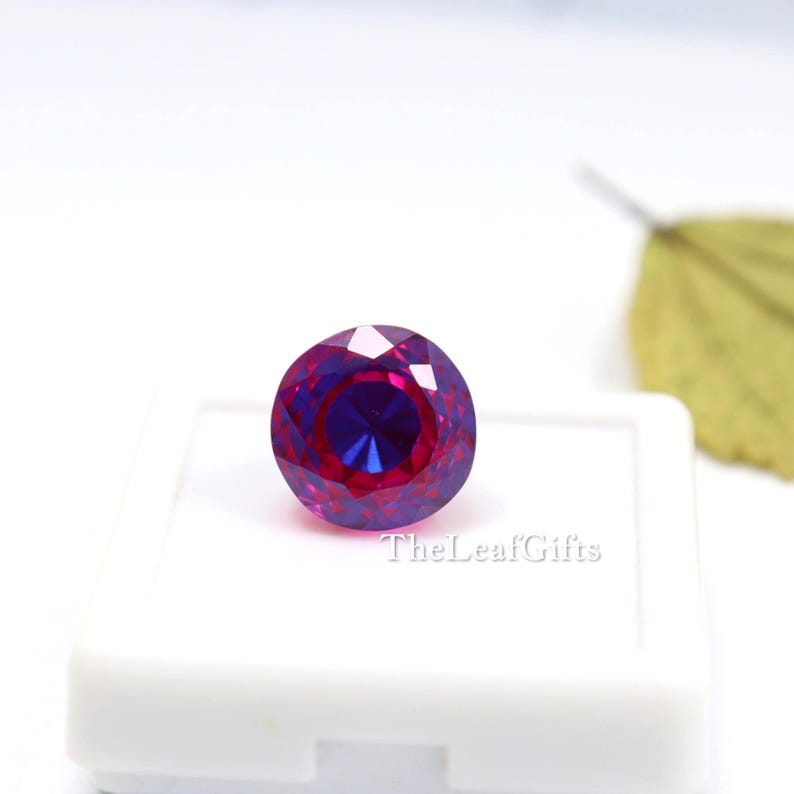 May include: A round, faceted gemstone with a colour gradient from deep magenta to royal blue. The gem is displayed in a white case, with the text "TheLeafGifts" visible.