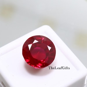 Pode incluir: Uma gema de rubi redonda e facetada. A gema é de cor vermelha profunda e possui múltiplas facetas que refletem a luz. A gema está sobre um fundo branco. O texto "TheLeafGifts" é visível no canto inferior direito.