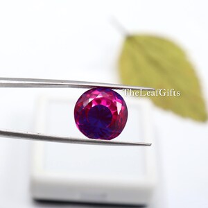 May include: A round, faceted gemstone held by tweezers. The gem has a vibrant colour gradient, transitioning from a deep magenta at the edges to a rich, dark purple in the centre. TheLeafGifts is written on the right side.