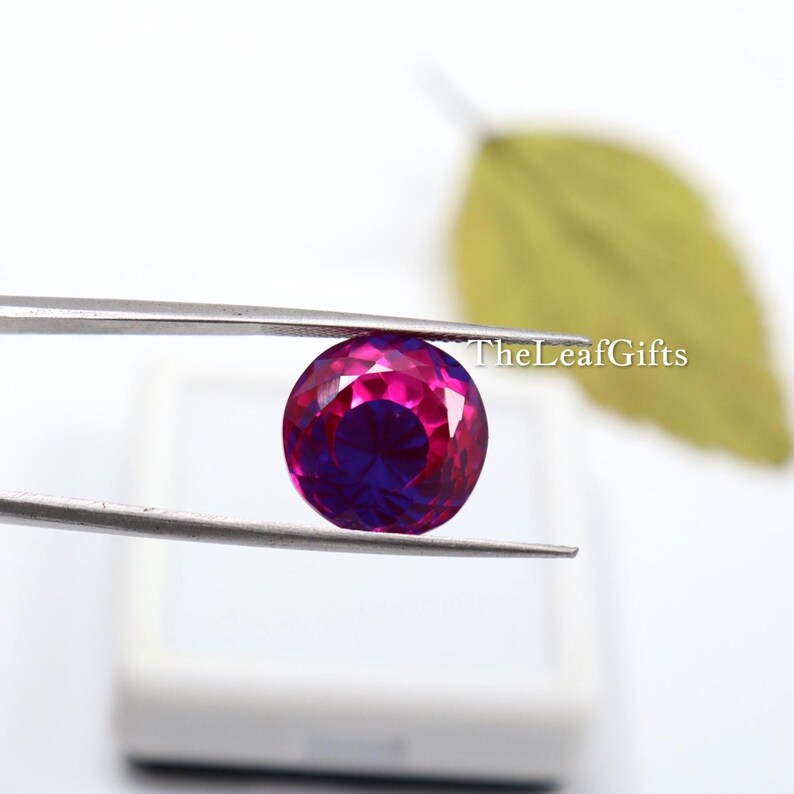May include: A round, faceted gemstone held by tweezers. The gem has a vibrant colour gradient, transitioning from a deep magenta at the edges to a rich, dark purple in the centre. TheLeafGifts is written on the right side.