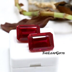 Pode incluir: Duas pedras preciosas retangulares, facetadas, de cor vermelha profunda. As gemas são exibidas em uma superfície branca, com o texto "THELEAFGIFTS" em preto. O fundo inclui um arranjo desfocado de elementos marrons e verdes.