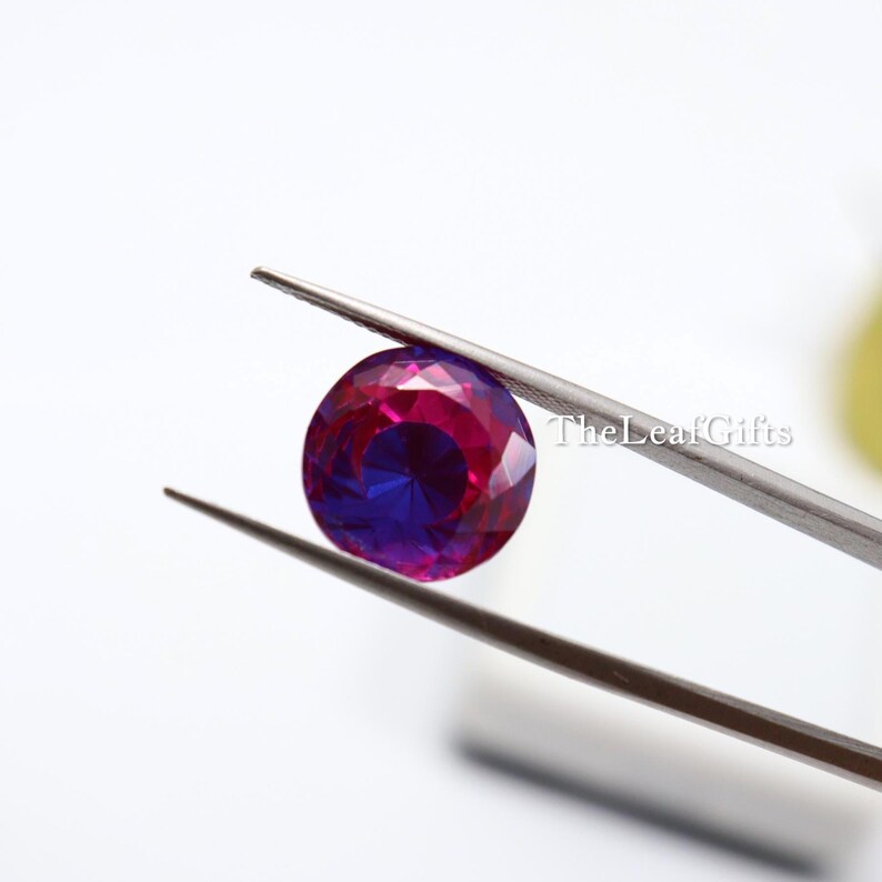 May include: A round, faceted gemstone held by tweezers. The gem displays a striking colour gradient, transitioning from a deep magenta at the edges to a vibrant blue in the centre. The Leaf Gifts is printed on the right side of the image.