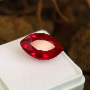 May include: A faceted, marquise-cut ruby gemstone. The gem is a vibrant red color with a lighter, oval-shaped interior. It rests on a white surface, with a blurred, natural background.