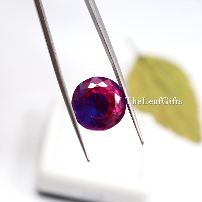 May include: A round, faceted gemstone held by tweezers. The gem has a deep magenta hue on the outer edges, transitioning to a rich, dark blue in the centre. TheLeafGifts is printed on the right side.
