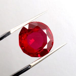 Peut inclure: Une pierre précieuse rouge foncé avec une taille facetée, tenue par des pinces sur un fond blanc.