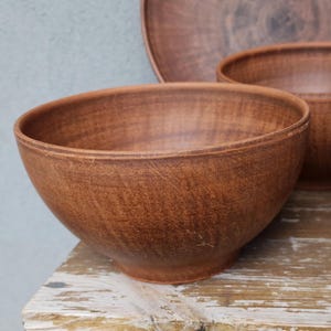 Può includere: Una collezione di ciotole e tazze in ceramica marrone fatte a mano. Le ciotole variano in dimensioni, con una grande ciotola ampia in primo piano. Le tazze hanno un aspetto rustico. La ceramica è esposta su una superficie di legno.