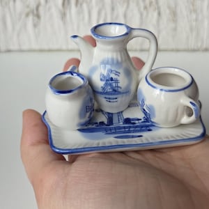 Könnte beinhalten: Miniatur-Teeservice aus weißer und blauer Keramik. Das Set umfasst eine kleine Teekanne, einen Sahnekännchen und eine Tasse, alle mit einem Windmühlen-Design verziert. Die Teile stehen auf einem passenden rechteckigen Tablett. Ein bezauberndes Sammlerstück.