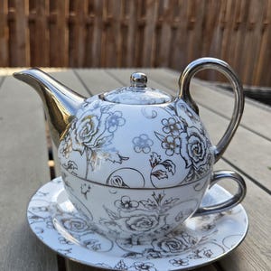 Puede incluir: Juego de té de porcelana blanca con diseño floral. El juego incluye una tetera con asa y boquilla plateadas, una taza de té y un platillo. El estampado floral presenta rosas y otras flores en plata y oro.