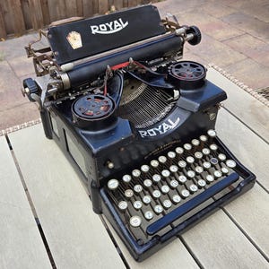 Puede incluir: Una máquina de escribir Royal negra con teclas blancas y la palabra "ROYAL" en blanco. La máquina tiene un carro negro y un marco de metal. Las teclas están dispuestas en QWERTY.