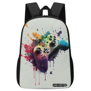 Può includere: Uno zaino nero con un pannello frontale bianco che presenta un design a schizzi di vernice colorato con un controller per videogiochi arcobaleno al centro. Il testo "Gamer Frenzy Wear" è stampato nella parte inferiore dello zaino.