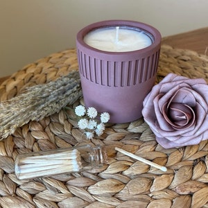 May include: A mauve candle in a textured ceramic jar with a white wick, a small glass jar of matches, a dried flower arrangement, and a mauve silk rose on a woven wicker surface.