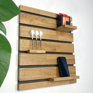 Op de afbeelding: Houten wandplank met een natuurlijke houtnerf afwerking. De plank heeft meerdere horizontale lamellen met kleine planken voor items zoals een smartphone, darts en kleine dozen. De achtergrond is donkergrijs.