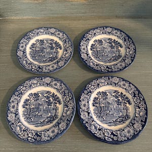 May include: Set of four blue and white ceramic plates with a floral border and a central image of a house with trees.