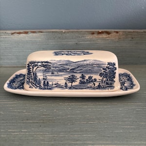 Può includere: Un burriera in ceramica color crema con un motivo blu a trasferimento. Il coperchio mostra una scena paesaggistica dettagliata con un lago, barche a vela, alberi e figure, mentre la base e la parte superiore sono decorate con motivi floreali.