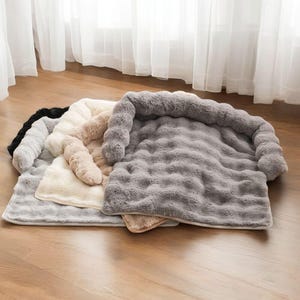 May include: A collection of plush pet beds in various neutral colors, including black, gray, cream, and tan. Each bed features a soft, textured surface and a raised, cushioned bolster for added comfort. The beds are rectangular and appear to be made of a faux fur material.