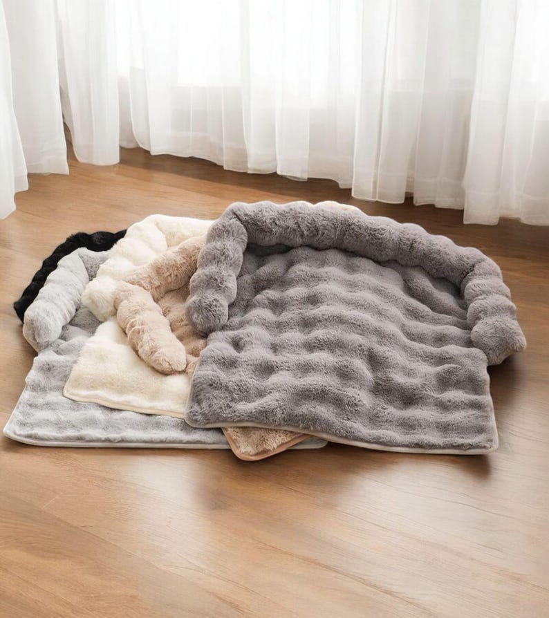 May include: A collection of plush pet beds in various neutral colors, including black, gray, cream, and tan. Each bed features a soft, textured surface and a raised, cushioned bolster for added comfort. The beds are rectangular and appear to be made of a faux fur material.