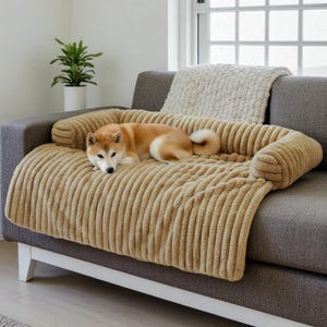 Op de afbeelding: Een bruin-witte Shiba Inu-hond rust op een beige, geribbeld dierenbed op een grijze bank. Het bed heeft opstaande randen en een zachte textuur. Een wit kussen en een potplant zijn zichtbaar op de achtergrond.
