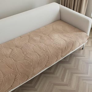 Könnte beinhalten: Ein beiges Sofa mit einem kissenbezug mit Blättermuster. Der Bezug hat ein erhabenes, strukturiertes Design aus großen, stilisierten Blättern. Das Sofa hat einen weißen Rahmen und steht auf einem hellen Holzboden.