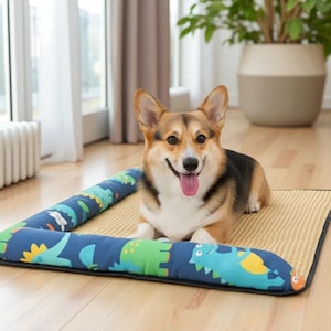 以下が含まれることがあります： 幸せなコーギー犬が長方形のペットベッドで休んでいます。ベッドはベージュ色のマットと、恐竜模様の青いボルスターが付いています。犬は茶色、黒、白の毛並みで、舌を出して笑顔です。