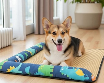 May include: A happy corgi dog rests on a rectangular pet bed. The bed has a tan mat and a blue bolster with a dinosaur pattern. The dog has a brown, black, and white coat and is smiling with its tongue out.