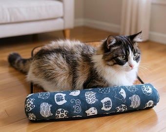 May include: A fluffy, multi-colored cat rests on a rectangular pet bed with a blue bolster pillow. The pillow is patterned with white cat faces and other shapes. The cat has brown, black, and white fur.