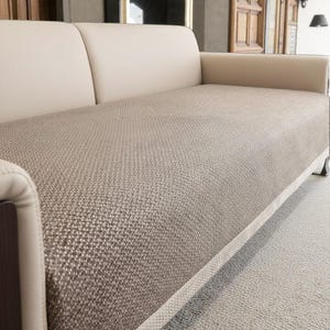 Puede incluir: Un sofá beige moderno con un cojín de asiento texturizado y tejido y respaldos y reposabrazos de cuero color crema. El sofá tiene un perfil bajo y se apoya en patas de metal plateado, ofreciendo una estética contemporánea.