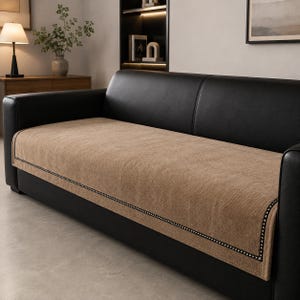 May include: A rectangular tan sofa cover with a textured weave pattern and a black and white geometric border trim. The cover is draped over the seat and front edge of a black leather sofa, providing a contrasting layer of fabric.