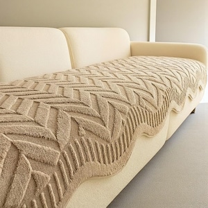 May include: A beige sofa with a tan, textured throw. The throw has a geometric pattern and a scalloped edge. The sofa has a curved back and armrests. The throw is draped over the sofa, creating a cosy look.