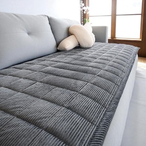 May include: A gray, corduroy-textured sofa cover with a quilted pattern. The cover is draped over a light gray sofa, with two matching back cushions and a decorative beige pillow. The sofa is near a window.