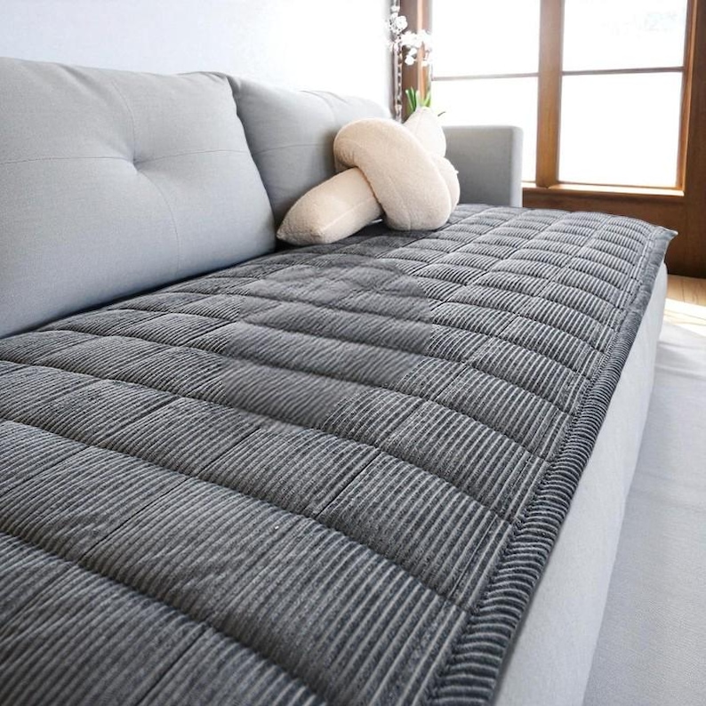 May include: A gray, corduroy-textured sofa cover with a quilted pattern. The cover is draped over a light gray sofa, with two matching back cushions and a decorative beige pillow. The sofa is near a window.