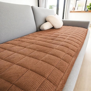 May include: A brown corduroy sofa cushion with a quilted pattern rests on a gray sofa. A pair of off-white decorative pillows are on the sofa. The cushion is rectangular and covers the seating area.