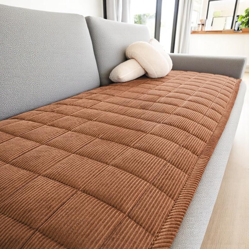 May include: A brown corduroy sofa cushion with a quilted pattern rests on a gray sofa. A pair of off-white decorative pillows are on the sofa. The cushion is rectangular and covers the seating area.