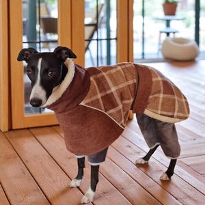 Op de afbeelding: Een zwart-witte hond met een bruine geruite jas met een witte fleece kraag en een grijze broek. De hond staat op een houten vloer. De jas heeft een bruin en wit ruitpatroon.