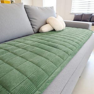 May include: A green corduroy sofa cushion cover with a quilted pattern. The cover is rectangular and covers the seat of a gray sofa. Two gray throw pillows and a white decorative knot pillow are on the sofa.