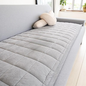 May include: A light gray, quilted sofa cushion with a corduroy texture, placed on a gray sofa. A cream-colored decorative pillow rests on the sofa. The cushion has a rectangular shape with a stitched grid pattern.