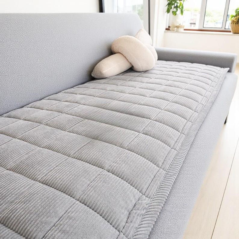 May include: A light gray, quilted sofa cushion with a corduroy texture, placed on a gray sofa. A cream-colored decorative pillow rests on the sofa. The cushion has a rectangular shape with a stitched grid pattern.