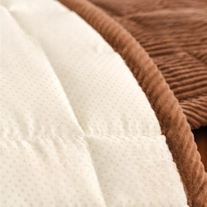 May include: Close-up of a quilted, off-white fabric with small, evenly spaced dots. The fabric is stitched in a diamond pattern. A brown, corduroy-like material borders the quilted fabric, creating a contrasting texture and color.