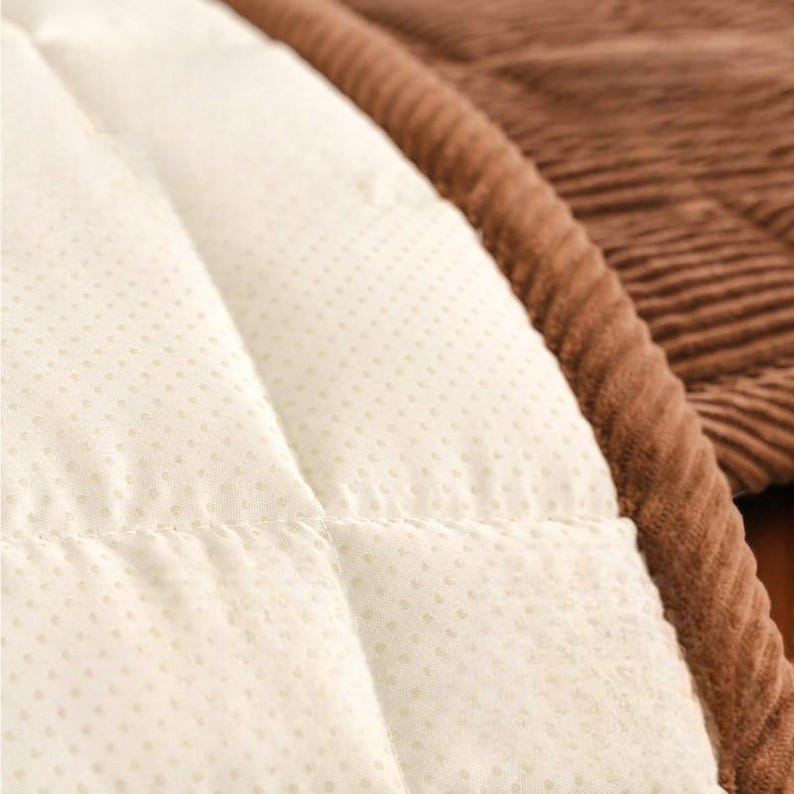 May include: Close-up of a quilted, off-white fabric with small, evenly spaced dots. The fabric is stitched in a diamond pattern. A brown, corduroy-like material borders the quilted fabric, creating a contrasting texture and color.