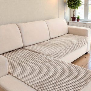 May include: A beige sectional sofa with a light grey textured throw. The sofa has a modern design and a neutral colour scheme, complementing the room's decor. The throw blanket adds comfort and style to the seating area.