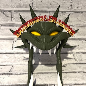Puede incluir: Un tocado verde con forma de dragón con ojos amarillos y dientes blancos. El tocado tiene púas y las palabras "DUNGEONS & DRAGONS" en rojo y amarillo. El tocado está hecho de un material duro y está sobre una pared de ladrillos blancos.