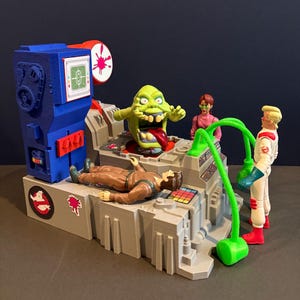 May include: A Ghostbusters playset featuring a green Slimer figure, a figure in a brown jumpsuit, and two Ghostbusters figures in white jumpsuits. The playset has a blue and gray color scheme with red accents and a Ghostbusters logo.