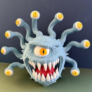 May include: A light blue spherical creature figurine with a textured surface, featuring a large central eye with a yellow iris, a wide mouth filled with sharp white teeth, a red interior, and ten long, wavy stalks topped with smaller yellow eyes.