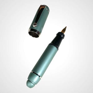 May include: A teal fountain pen with a gold nib and a black grip. The cap is off, revealing the pen's design. The pen has silver accents and a rounded tip. The pen is set against a white background.