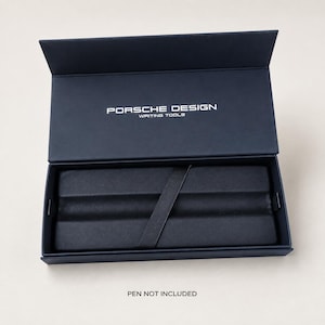May include: A dark navy blue rectangular presentation box with a hinged lid. The interior features a black velvet-lined tray with a diagonal fabric strap. The text PORSCHE DESIGN WRITING TOOLS is printed in white on the inside of the lid.