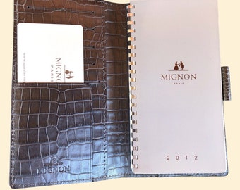 May include: A brown leather bound planner with a gold coil binding. The cover has a crocodile embossed pattern and the inside cover has a pocket for business cards. The planner is open to a blank page with the year 2012 printed at the bottom. The planner has the Mignon Paris logo on the cover and the inside page.