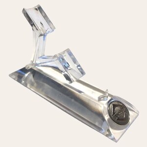 May include: A clear acrylic display stand with a rectangular base and an angled support structure. The stand features a small, circular emblem on the base. The design is modern and minimalist, suitable for showcasing collectibles or small items.