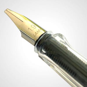 May include: Close-up of a fountain pen nib with the text "1.9 LAMY" engraved on the gold-colored tip. The pen's body is clear, revealing the internal ink mechanism. The nib is angled towards the upper left.